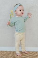 Baby wearing the Sage Leaf top and ribbed leggings, standing against a wall – showcasing a clean round-neck design and contrasting trim detail
